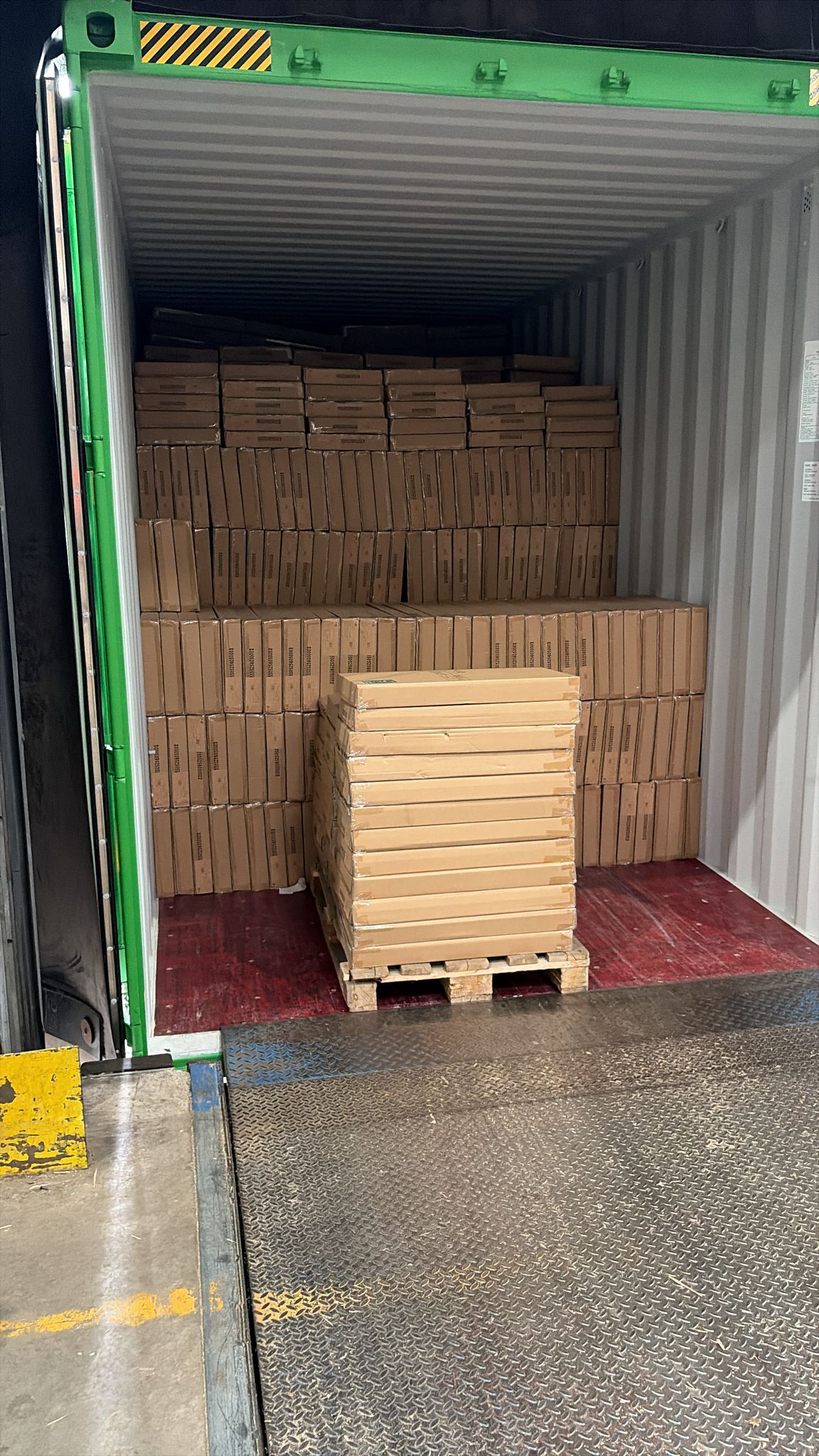 Een geopende zeecontainer met groene deuren aan een laaddok, half gelost: achterin liggen de platte dozen van de flatpacks nog in rijen opgestapeld, vooraan op de containervloer staat één pallet vol gestapelde flatpacks klaar.